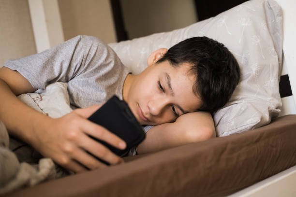 A teenage boy lying in bed and looking at his smartphone.